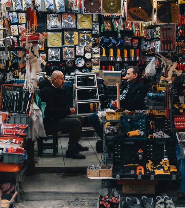 Two men in conversation at a bustling street market stall filled with tools and accessories.